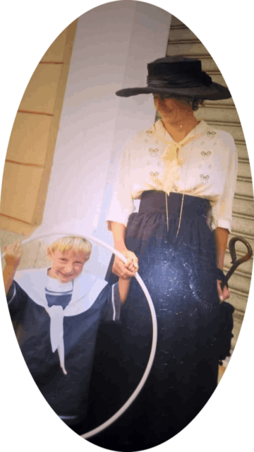 Ritratto Ovale Luciana B&B Corsica - Luciana Masinari, la padrona di casa, in abbigliamento ottocentesco, gonna blu, camicetta beige ricamata e ampio cappello blu, in compagnia del giovane figlio vestito alla marinara.